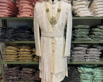 Op de afbeelding: Een witte geborduurde mannenoutfit met een bijpassende witte tulband. De outfit heeft een lange, vloeiende jas met een parel- en groene kralenketting. De outfit wordt getoond op een mannequin in een winkel.