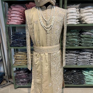 May include: A gold embroidered men's sherwani with a matching turban. The sherwani has a long, flowing design with intricate embroidery and a pearl necklace.