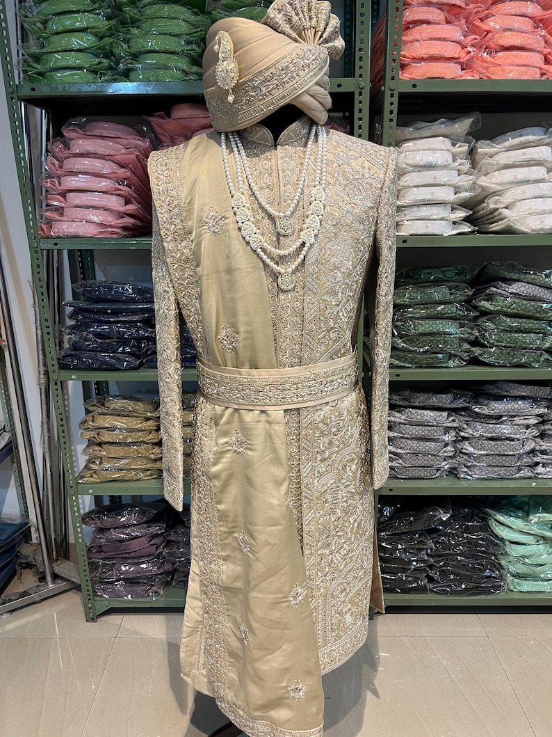 May include: A gold embroidered men's sherwani with a matching turban. The sherwani has a long, flowing design with intricate embroidery and a pearl necklace.