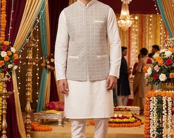 May include: A man wearing a white kurta pajama set with a light gray patterned vest. The outfit is worn with matching shoes. The background features a decorated event space with floral arrangements and decorative lighting.