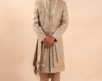 Puede incluir: Un traje sherwani beige bordado con pantalones y zapatos a juego. El atuendo incluye un turbante con una pluma y un collar de perlas. La chaqueta de manga larga presenta intrincados bordados dorados y un diseño plisado.