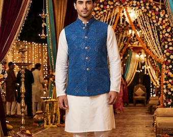 Könnte beinhalten: Ein Mann in einem weißen Kurta und Hose, kombiniert mit einer blauen bestickten Weste mit goldenen Knöpfen. Das Outfit ist für eine formelle Veranstaltung geeignet. Der Hintergrund zeigt dekorative Blumenarrangements und Kronleuchter.