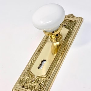 May include: A door knob set featuring a smooth, round white porcelain handle mounted on a polished brass rectangular backplate. The backplate displays an intricate embossed border pattern and a central keyhole opening for a mortise lock mechanism.