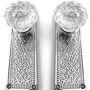 May include: Two vintage door handles. Each handle features a clear, faceted glass knob atop a decorative, rectangular metal plate. The plates have an ornate, embossed floral pattern. The hardware is in a silver tone, and the background is white.