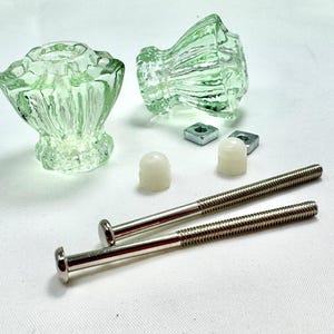 May include: Two light green glass cabinet knobs with a textured, fan-like design. The knobs are accompanied by silver screws, white plastic caps, and square metal nuts. These decorative knobs are ideal for furniture restoration projects.