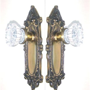 May include: Two door knobs with clear, faceted glass handles and brass-toned metal bases. Each knob is attached to an elongated, ornate rectangular backplate featuring dark, aged metal detailing and a smooth, central brass-colored oval panel.