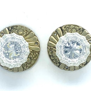 May include: Two circular cabinet knobs featuring a clear, faceted glass center with a scalloped edge. Each knob is mounted on a round, antique brass-toned metal backplate decorated with embossed floral patterns and radiating fan-like lines.