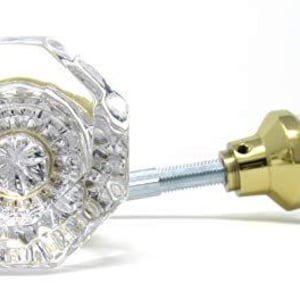 May include: Two clear, faceted glass cabinet knobs with an octagonal shape. Each knob features a polished gold-tone metal base and a central circular design with radiating patterns. A threaded metal bolt extends from the back of the knobs.
