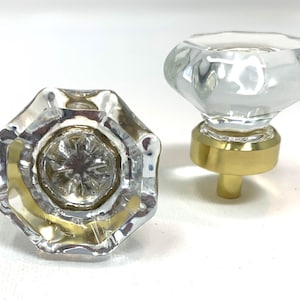 May include: Two clear glass cabinet knobs with a faceted design. The knobs have a gold-colored base and stem. One knob is viewed from the top, and the other is viewed from the side. These knobs are suitable for furniture restoration or adding a vintage touch.