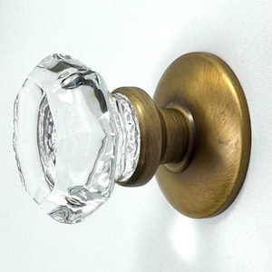 May include: A clear, faceted glass knob attached to a circular, brushed brass metal base. The knob features a multi-sided geometric design that reflects light, while the mounting plate has a smooth, matte finish in a warm metallic tone.