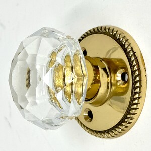 May include: A clear, multi-faceted glass door knob attached to a circular, polished brass backplate. The backplate features a decorative rope-style edge and two visible screw holes for mounting, showcasing a metallic gold finish throughout.