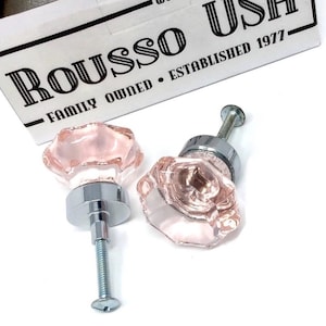 May include: Two pink glass cabinet knobs with a faceted design and chrome bases. The knobs are next to a white box with the text "ROUSSO USH FAMILY OWNED ESTABLISHED 1977".