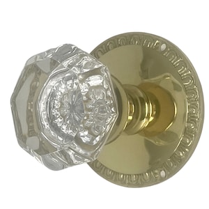 May include: A clear, faceted crystal door knob with a brass-colored base and stem. The round base features an ornate, repeating pattern around the edge. The knob is a classic design, suitable for interior doors.