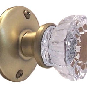 May include: A door knob featuring a clear, faceted glass handle with a multi-sided design. The base is a circular, brushed brass-toned metal plate with two visible screws, connecting to a matching brass-toned cylindrical neck and collar.