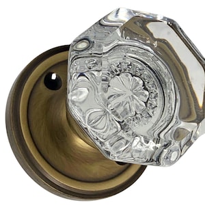 May include: A clear, faceted glass door knob with an octagonal shape. The knob is mounted on a round, antique brass-colored backplate. The glass knob has a decorative, floral-like pattern.