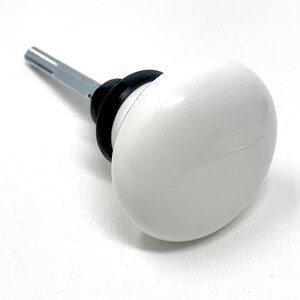 May include: A white, oval-shaped cabinet knob with a glossy finish. The knob is attached to a silver metal stem with a black ring at the base. The knob is designed for use on drawers, cabinets, or furniture.