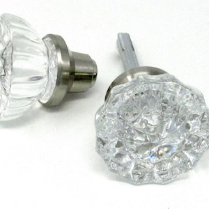 May include: Two clear glass cabinet knobs with a faceted design. The knobs have a silver-colored metal base and stem. The knobs are designed for use on cabinets, drawers, or furniture.