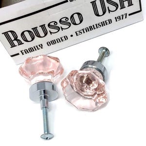 May include: Two pink crystal cabinet knobs with silver hardware. The knobs have a faceted, flower-like design. The box in the background reads "Rousso Ush Family Owned Established 1977."