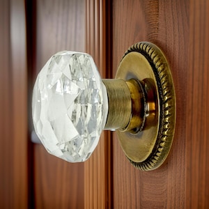 May include: A clear, multifaceted glass door knob with a geometric, diamond-like shape. The knob is attached to a circular, antique brass-toned metal backplate featuring a decorative rope-style border, mounted onto a vertical wooden door panel.
