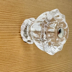 May include: A clear, faceted glass cabinet knob with a multi-sided, flower-like shape and a smooth, rounded profile. The center of the knob features a small, circular, polished silver-tone metal screw cap that secures the piece to the surface.