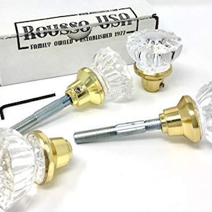 May include: Three clear, faceted glass cabinet knobs with gold-toned metal bases and threaded metal spindles. A white cardboard box labeled Rousso USA Family Owned Established 1977 is visible, along with a small black L-shaped hex key tool.