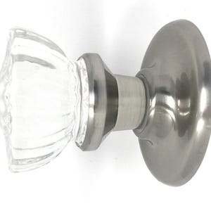 May include: A door knob featuring a clear, faceted glass handle with a fluted design. The handle is attached to a circular, brushed nickel metal base plate and a matching cylindrical neck, providing a functional hardware component for doors.
