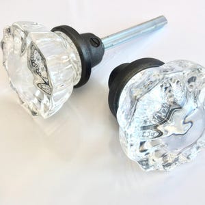 May include: Two clear glass door knobs with a faceted, multi-sided design. Each knob features a dark, matte-finished metal base and a central metal spindle extending from the back, designed for installation on a door or cabinet surface.