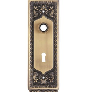 May include: A rectangular brass door plate with an antique finish, featuring an ornate border with scrollwork and floral motifs. The plate includes a circular opening for a knob at the top and a keyhole opening below, with two screw holes.