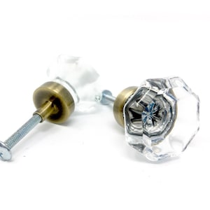 May include: Two clear, faceted glass cabinet knobs with antique brass-colored bases and silver screws. The knobs are designed with an octagonal shape, reflecting light. The hardware is set against a white background.