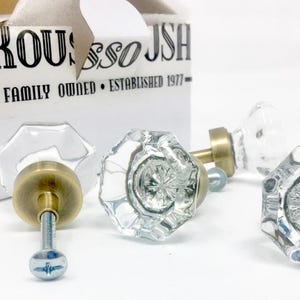 May include: Clear glass and brass cabinet knobs with a faceted design are displayed near a white box with the text "ROUSSO JSH - FAMILY OWNED - ESTABLISHED 1977-". The knobs have a vintage aesthetic.