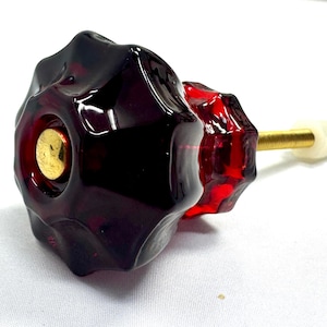May include: A deep red, translucent glass cabinet knob with a faceted, star-like shape and a smooth, rounded surface. The center features a circular gold-toned metal inlay. A threaded brass-colored metal bolt extends from the back with a white nut.