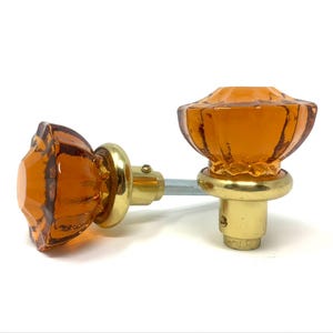 May include: Two amber-colored glass door knobs with a faceted design. The knobs have a brass base and are connected by a silver-colored rod. The glass has a warm, orange hue, and the knobs are set against a white background.