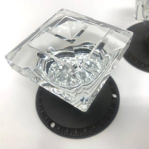 May include: Clear glass door knobs with a diamond-shaped design. The knobs are mounted on black metal plates with decorative edges. The glass has a faceted appearance, reflecting light. Hardware for doors.