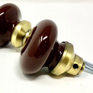 May include: Pair of dark brown ceramic door knobs with a glossy finish. The knobs have a rounded shape and are mounted on brass-colored metal bases. The hardware includes a threaded rod for installation.