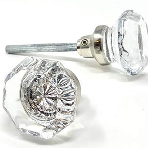 May include: Two clear, faceted glass cabinet knobs with silver-toned metal hardware. One knob is lying down, revealing its intricate interior design. The other knob is upright, attached to a long metal screw.