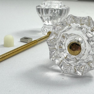 May include: Two clear, faceted glass cabinet knobs with an octagonal shape and a central gold-toned metal insert. A long, threaded gold-toned metal bolt, a small square metal nut, and a white plastic cap are included with the hardware.