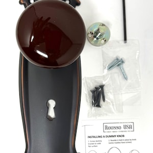 May include: A dark brown ceramic door knob mounted on an oil-rubbed bronze backplate with a keyhole. Included are a metal mounting bracket, an Allen wrench, two sets of screws in plastic bags, and an instruction sheet titled INSTALLING A DUMMY KNOB.