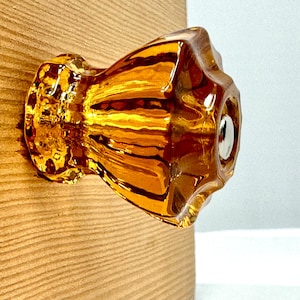 May include: A translucent amber-colored glass cabinet knob with a faceted, multi-sided design and a central silver-toned metal screw cap. The knob features a fluted, star-like shape that catches the light, creating highlights and shadows.