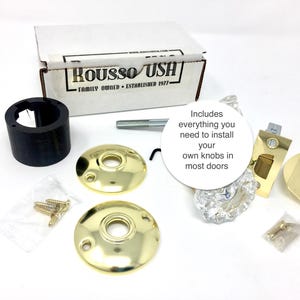 May include: A door knob installation kit with various components, including gold-colored rosettes, a clear crystal knob, and a black cylindrical piece. The kit includes everything needed to install door knobs. The box reads "ROUSSO USH FAMILY OWNED ESTABLISHED 1977-"