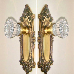 May include: A pair of door knobs featuring clear, faceted glass handles attached to ornate, rectangular brass backplates. The metal plates display a dark, aged finish with intricate embossed detailing surrounding the central handle mechanism.