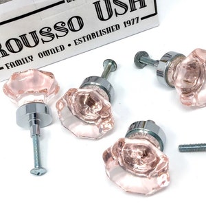 May include: Four pink glass cabinet knobs with chrome bases and screws. The knobs have a faceted design and are in a box labeled "Rouso USA Family Owned - Established 1977".