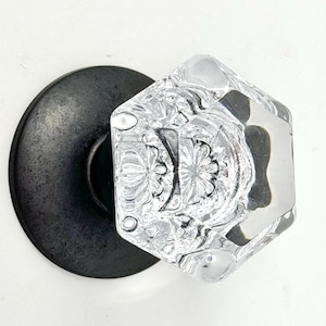 May include: Clear, faceted glass door knob with a floral design. The knob is mounted on a round, black metal backplate. The glass knob has a diamond shape and reflects light.