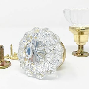 May include: Two clear glass cabinet knobs with a faceted, multi-sided design and gold-toned metal bases. One knob is shown from the front, while the other is upright. Included are two gold-toned metal mounting plates with threaded stems and screws.