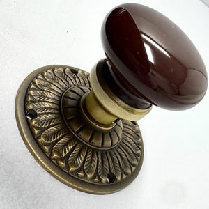 May include: A brown oval door knob with a brass base and a decorative leaf pattern. The knob has a glossy finish and is mounted on a round backplate. The hardware is designed for a door.