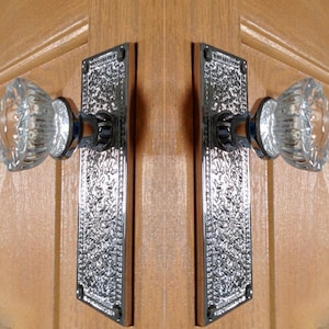 May include: A pair of door handles featuring clear, faceted glass knobs attached to rectangular, polished chrome backplates with an intricate, embossed floral pattern. The hardware is mounted onto a light brown, wood-grained surface.