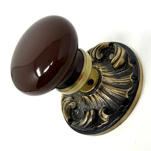 May include: A round, glossy dark brown ceramic door knob attached to a circular metal backplate. The backplate features an ornate, raised relief pattern with a dark, weathered finish and polished brass-toned highlights along the edges.