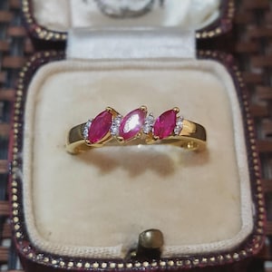 Puede incluir: Un anillo de oro con tres piedras preciosas rojas de corte marquesa dispuestas horizontalmente. Pequeñas piedras de acento redondas y transparentes están engastadas entre cada una de las piedras rojas, creando una banda decorativa continua.
