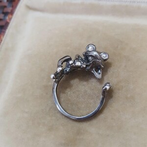 May include: A silver ring with a detailed frog design. The frog is positioned on the ring band with its mouth open. The ring has a polished finish. The ring is set against a neutral background.