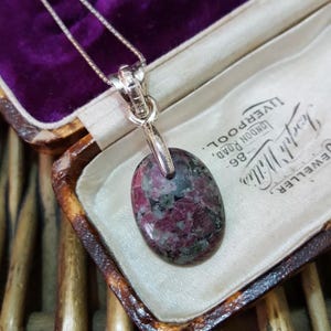 May include: A silver chain necklace with a pendant featuring a pink and green oval-shaped gemstone. The pendant is set in a silver bail and hangs from a silver chain. The necklace is displayed in a velvet-lined jewellery box.
