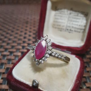 Puede incluir: Un anillo vintage con una piedra central de rubí en forma de marquesa, rodeada por un halo de pequeños diamantes redondos. El anillo está engastado en una banda de plata y se presenta en un joyero de terciopelo rojo. El diseño del anillo es clásico y elegante.